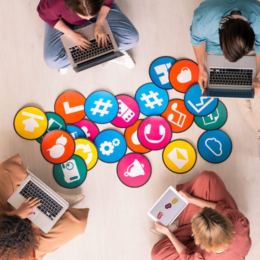 Top view of four young friendly people in casualwear sitting by multi-color papers with icons on the floor and using mobile gadgets